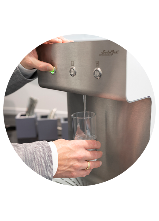 Eine Person füllt ein Glas mit Wasser an einem Edelstahl-Spender nach, im Hintergrund stehen Besteckbehälter.